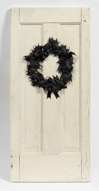 Circular faux ivy wreath with artificial holly and black bow hung on distressed white wooden door
