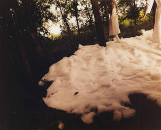 Two partially obscured figures in white dresses standing on a layer of cotton in wooded landscape