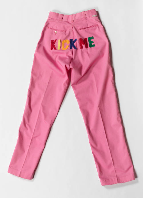 Pink pants, laying flat to display backside, with rainbow letters spelling “KICK ME” across the seat