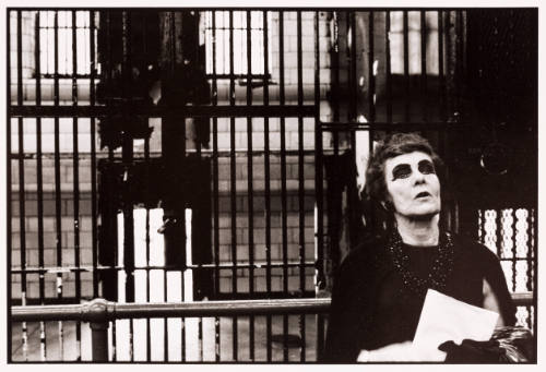 A person with dark eye make-up and necklace sits against a railing in a barred industrial interior