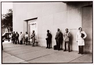 A line of evenly spaced people people in coats standing on a wide sidewalk against a building wall