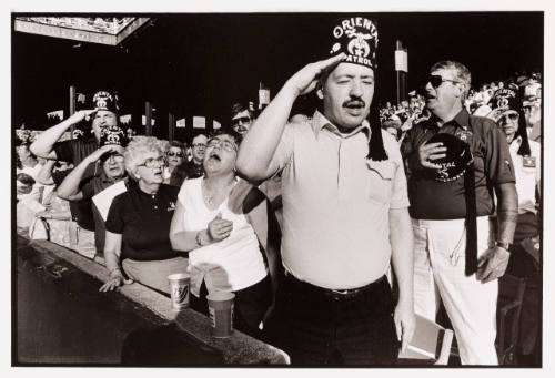 A crowd of Shriners in a stadium who are standing, saluting, or with hand over hearts