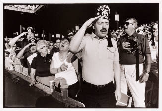 A crowd of Shriners in a stadium who are standing, saluting, or with hand over hearts