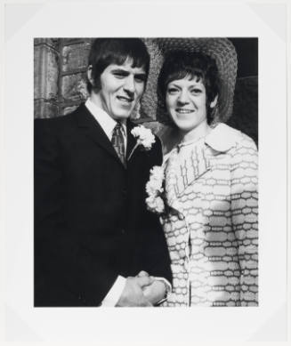 Smiling man in dark suit with boutonnière stands next to smiling woman in hat and coat with corsage