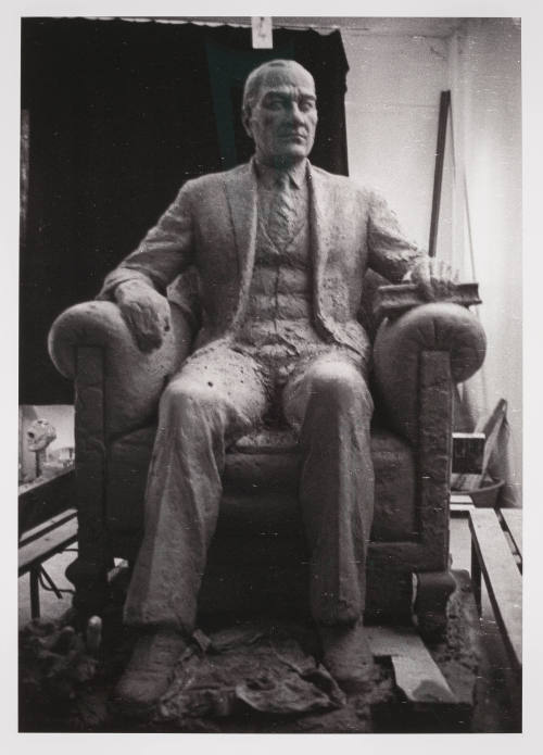 A black-and-white photograph of a sculpture of a man in a suit sitting in an armchair