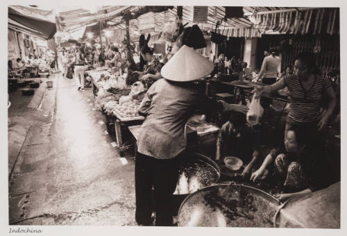 Photo of person in foreground with conical hat reaching for bag from vendor in busy marketplace