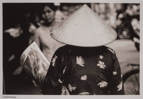 Black-and-white photo with person seen from behind with conical hat and street scene in background