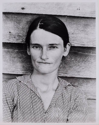 Portrait of a female-presenting figure with dark, drawn-back hair in front of a clapboard structure