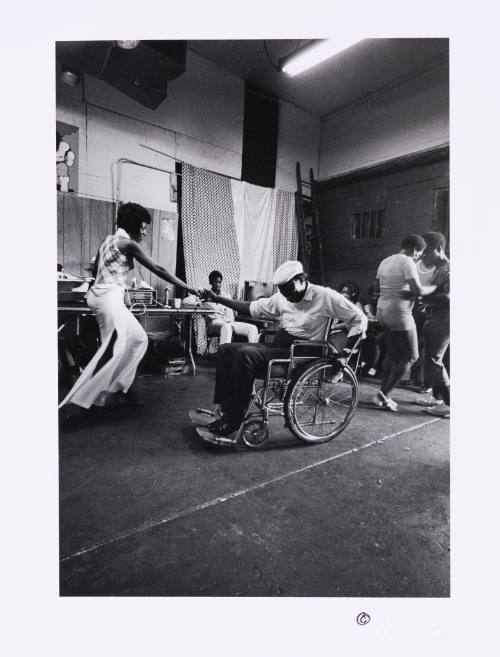 A Black man in a wheelchair smiles and dances with a standing Black woman wearing white bellbottoms