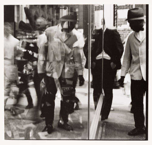 Man in jacket and fedora stands on a busy sidewalk and peers into store window with reflections