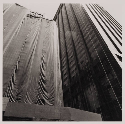 Tight black and white photograph looking upward at two skyscrapers, one scaffolded and draped in clo