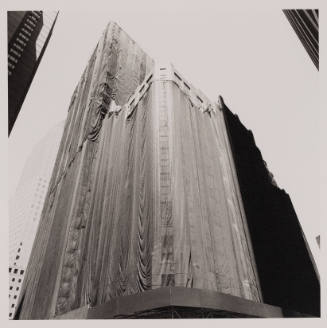 Tight black and white photograph looking upward at two skyscrapers, one scaffolded and draped in clo