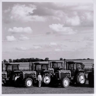 Landscape with puffy clouds, in the foreground the artist is posed in front of a line of tractors