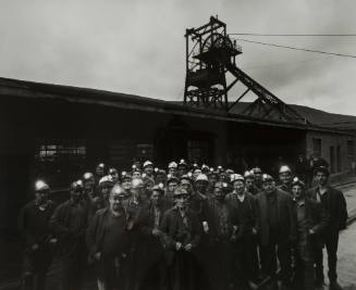 A group of people wearing miner’s caps face the camera while standing in front of a mining shed