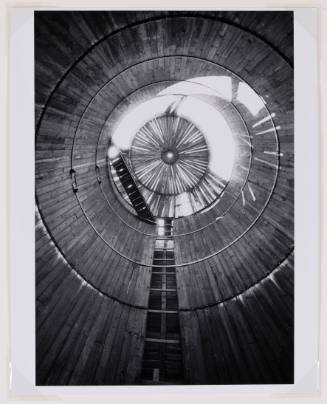 Photo taken from inside an empty silo looking up, showing wood paneling and a walkway across the top