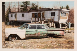 Vintage rusted, green car in front of a white and beige mobile home with trees in background