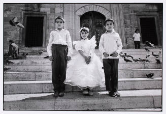 Two boys in long pants and long sleeve shirts flank a girl who is wearing a dress with big ruffles