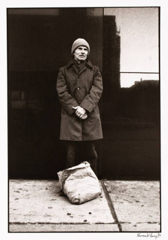 Black-and-white photo of a man standing near a building on a street wearing a coat and knit cap