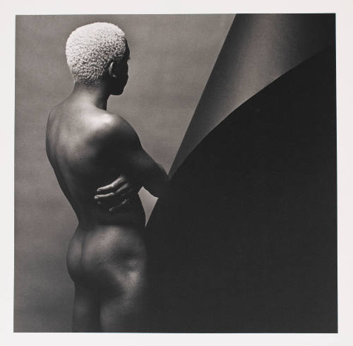 Nude person with dark skin tone and bleached hair seen from the back with their arms crossed
