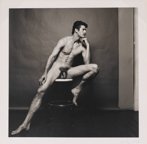 Portrait of light-skinned nude man seated on stool with legs open, in front of paper backdrop
