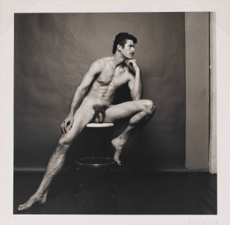 Portrait of light-skinned nude man seated on stool with legs open, in front of paper backdrop