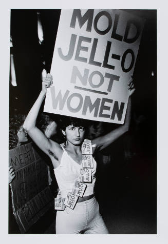 Black-and-white photograph of woman holding up sign that reads "MOLD JELL-O NOT WOMEN" 