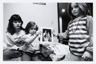 Black-and-white photo of light-skinned woman and two children holding photo of themselves with a man