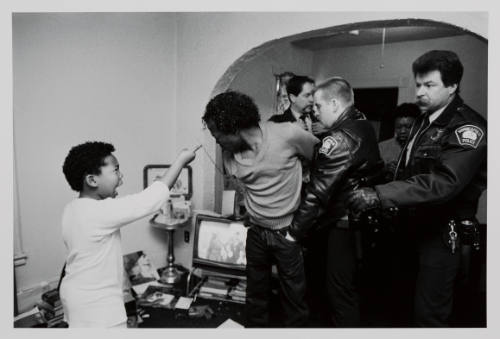 Black-and-white photograph of Black man held by white police officer while Black child yells at him
