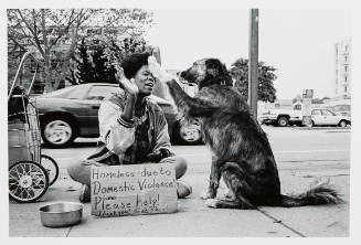 Black woman sitting on sidewalk high-fiving a long haired dog and holding a sign asking for help