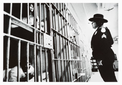 Female-presenting guard stands outside a prison cell and looks at two women on bunks inside