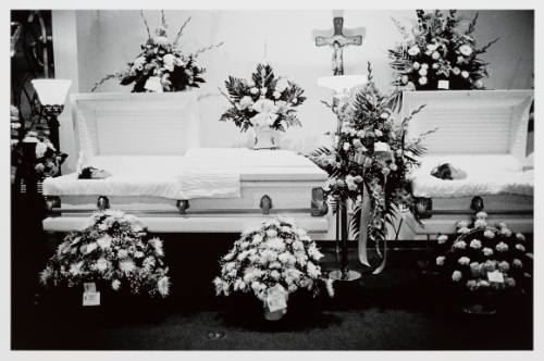Interior of funeral home with two opened white caskets surrounded by seven bouquets of flowers