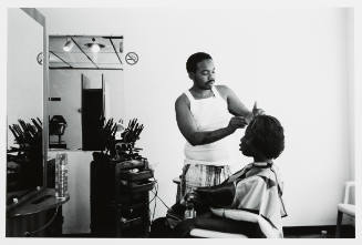 Photograph of two people with dark skin tone in a salon, a man cuts his client’s short hair