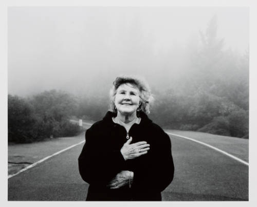 Standing in the middle of a foggy road, a light-skinned older woman places her hand over her heart 
