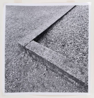 Black-and-white close-up photograph of the corner of a curb with gravel on both sides