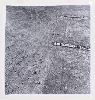 Square, black-and-white photograph of the ground with dirt and pebbles and remnants of concrete path