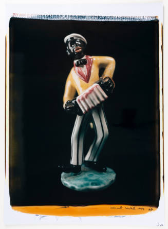 Color photo of figurine caricaturing a Black man in yellow suitcoat, striped pants playing accordion