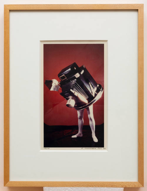 Color photograph of a vintage camera with human legs pointed down against a red backdrop